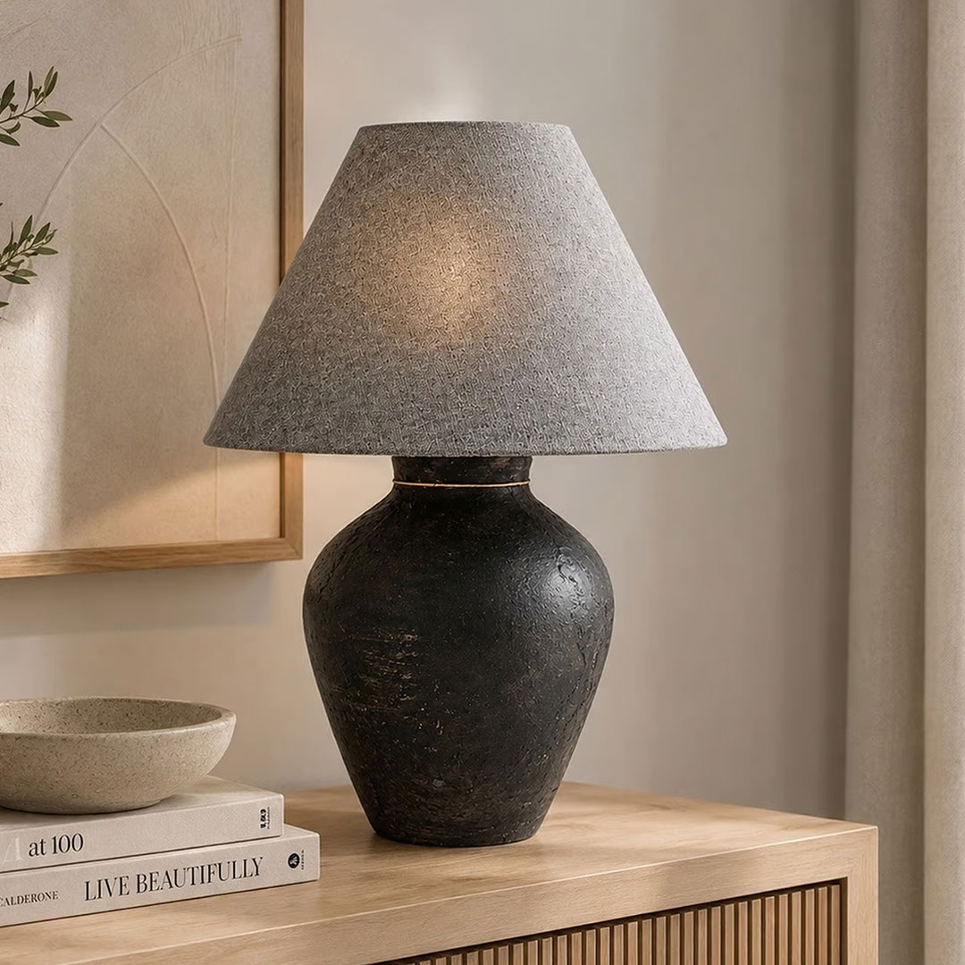 - A table lamp with a dark, rounded ceramic or stone-like base and a light gray fabric shade. 
- Appears suitable for a unisex/neutral decor style, likely intended for living room or bedroom use. 
- Visible features include a matte charcoal base with a subtle texture and a classic tapered-gray lampshade.